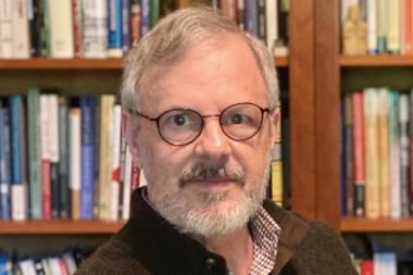 A gray haired white man wearing glasses sits in front of a bookshelf